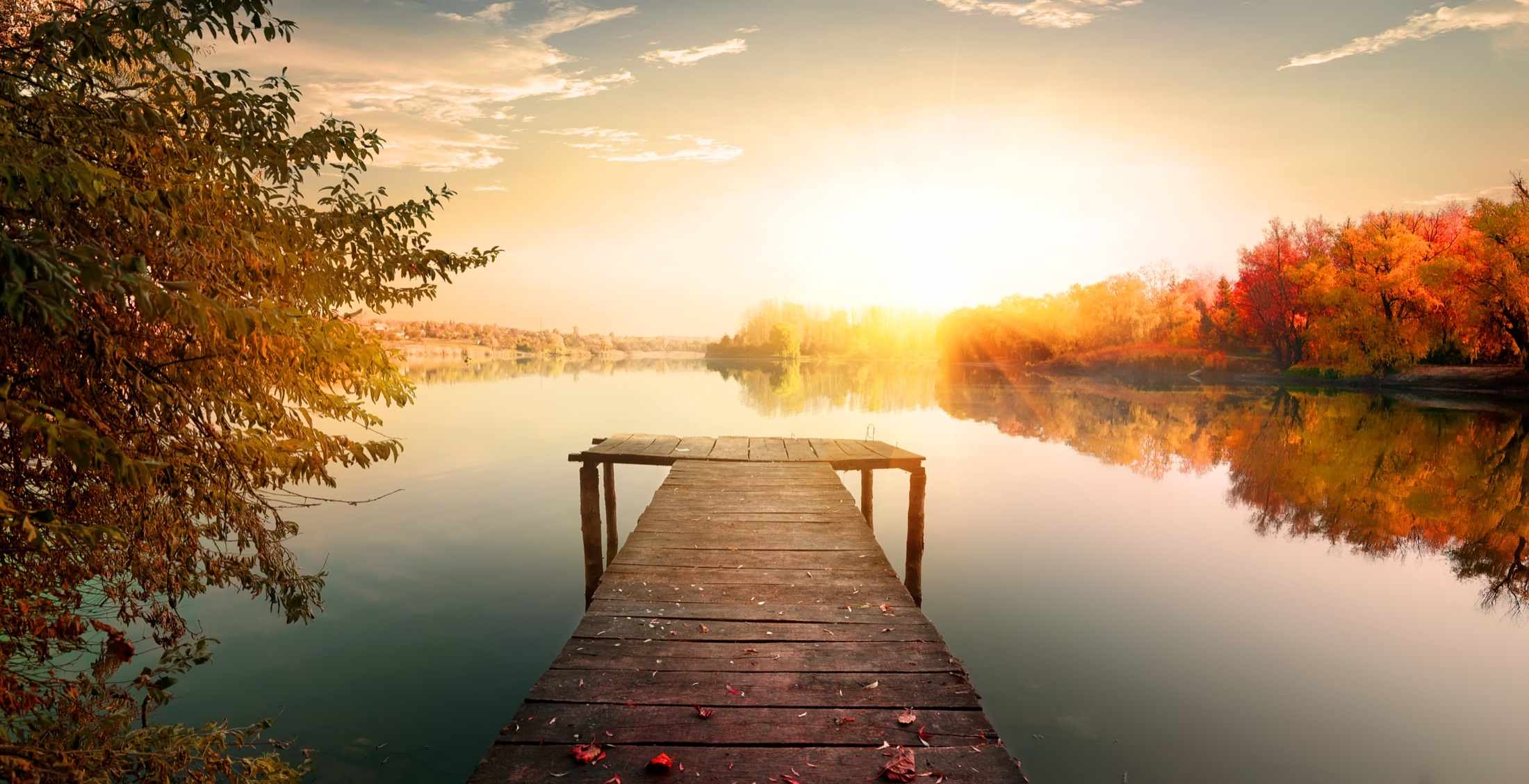A wooden dock reaching into a still lake at golden hour, autumn trees glowing on the far bank.